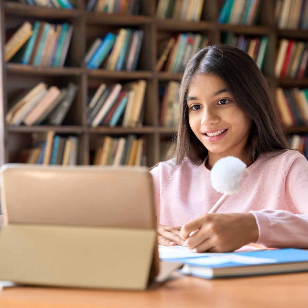 Happy smiling arab indian girl student watching learning online video class webinar in virtual classroom on digital tablet device. Kid school girl writing on table at home. Study from home.